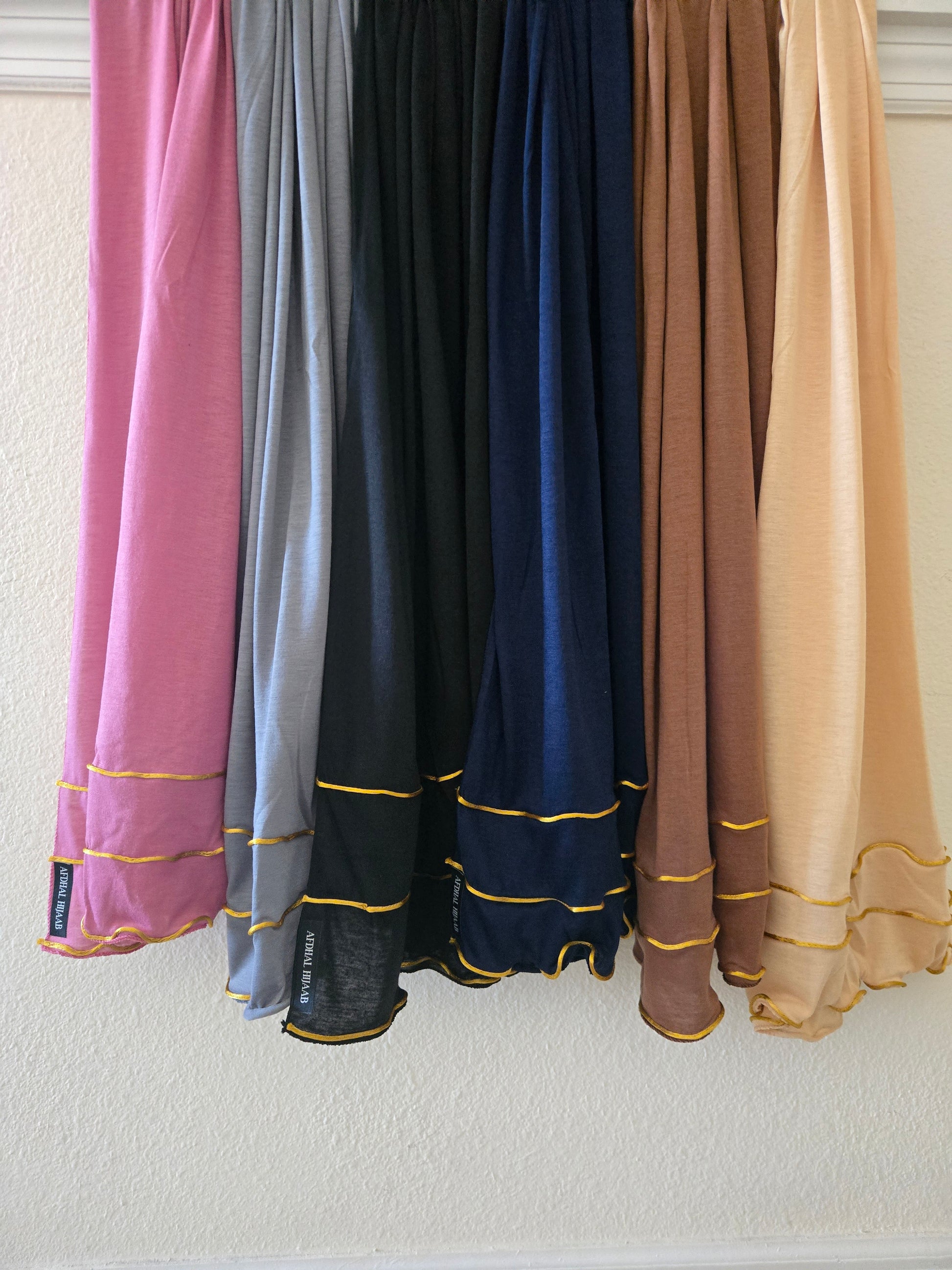 Set of colorful cotton jersey hijabs with gold trim hanging against a neutral wall.